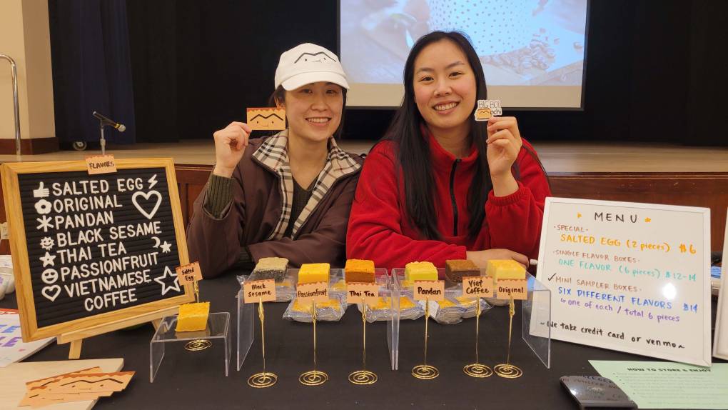 The two founders of Big Boi Mochi hold up small cards showing the logo for the Asian American home bakery business, with a tray of their butter mochi in front of them.