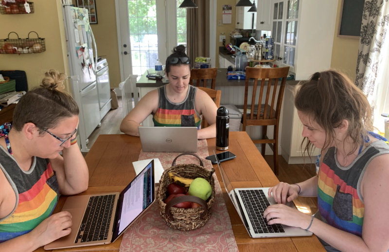 Three women sit around a rectangular kitchen table, each working on individual laptops. They are all wearing sleeveless tank tops with rainbow colors on the front.