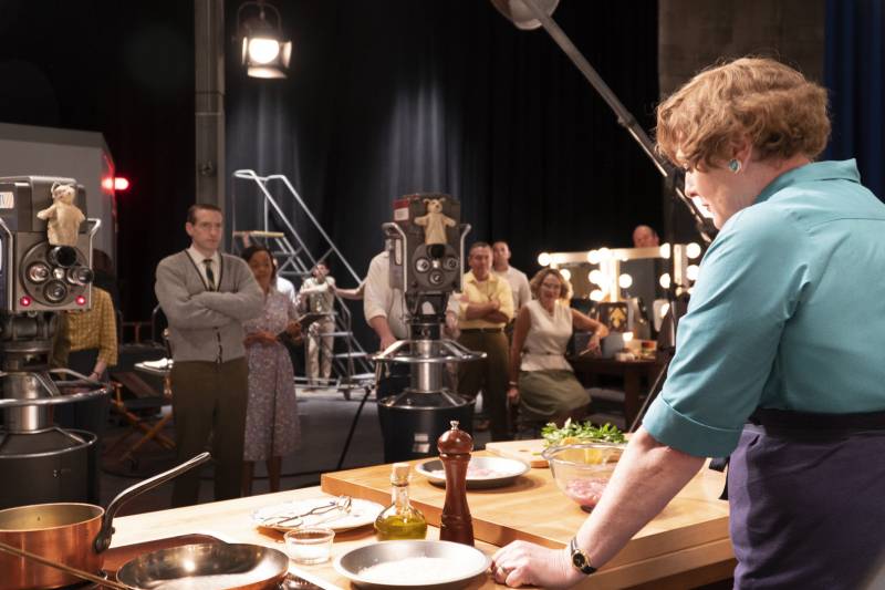 A woman stands behind a counter authoritatively. Before her is a counter covered in cooking equipment, two TV cameras trained on her and eight men and women avidly watching her. 