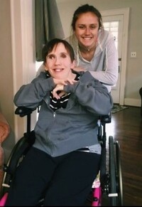 A smiling middle-aged woman sits in a wheelchair, as a teenage girl stands behind her, leaning on the wheelchair and smiling broadly.