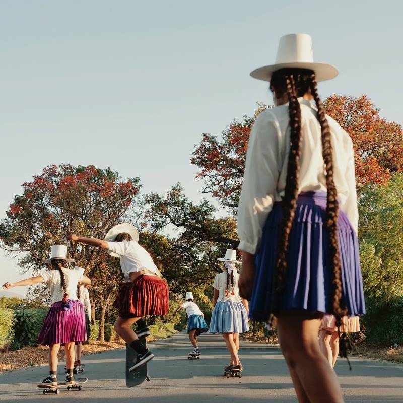 Girls in traditional Bolivian attire, wearing their hair in long braids, skateboard together in a park, trees in the distance.