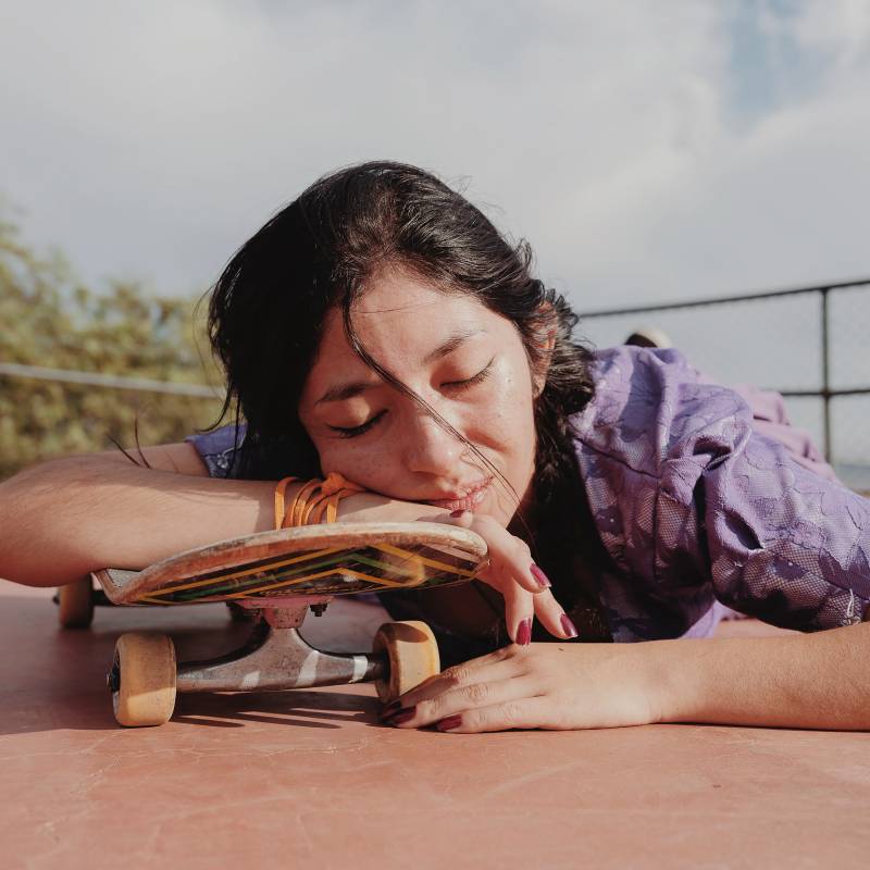 A young woman with closed eyes rests her right arm and head on a skateboard deck, as she lies on the ground, a serene expression on her face.