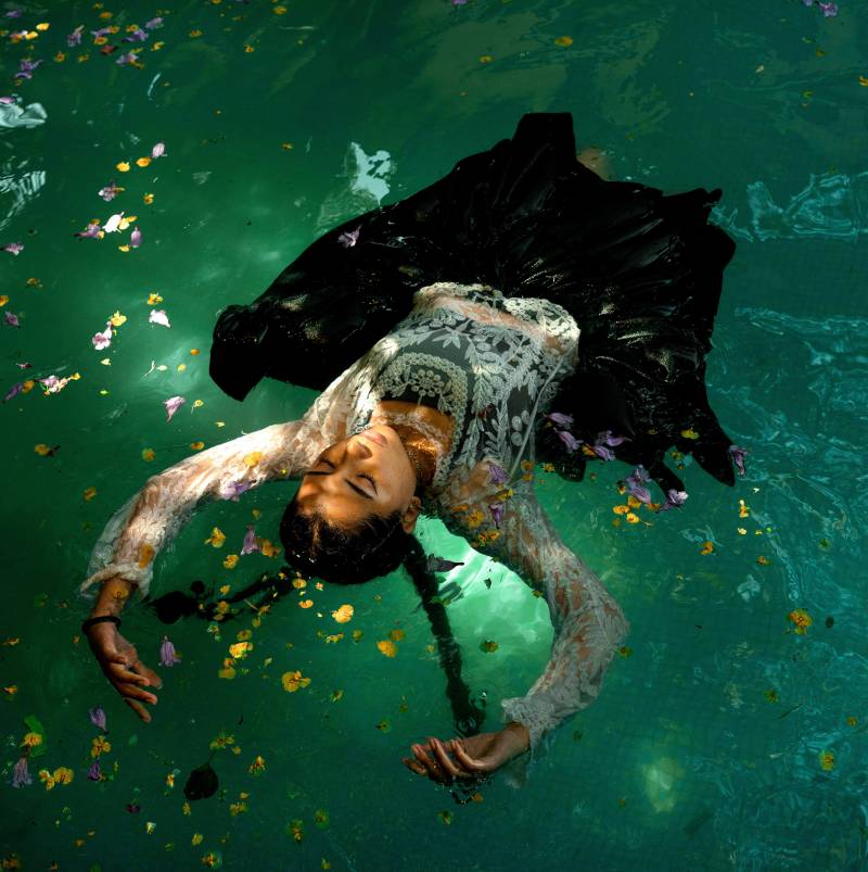 A young woman floats on her back in green water, her dress and braids floating around her.