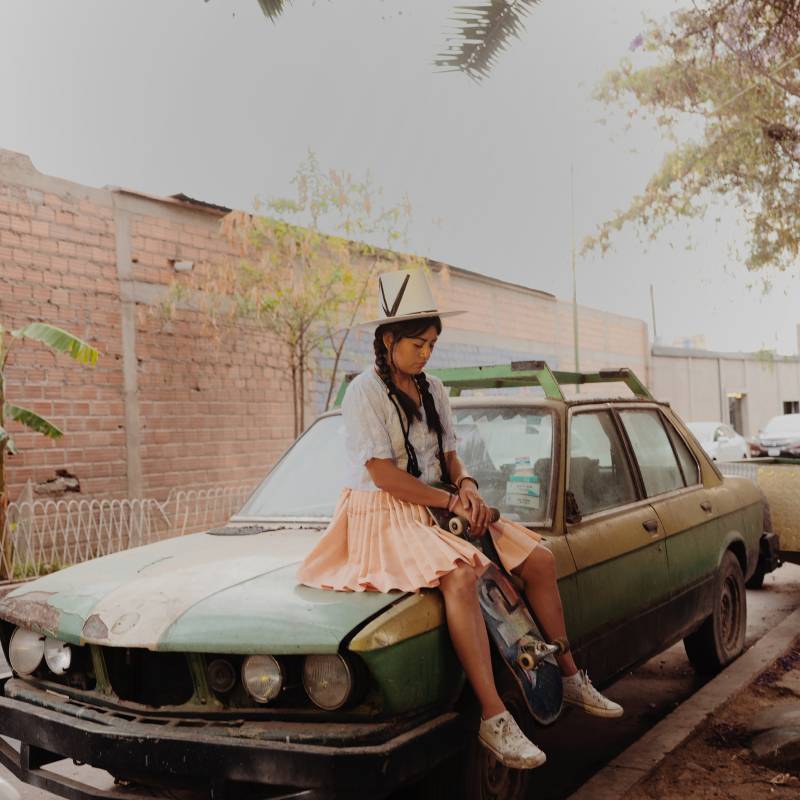 A young woman in traditional Bolivian hat, skirt and braids sits on a dusty car, skateboard resting between her legs.