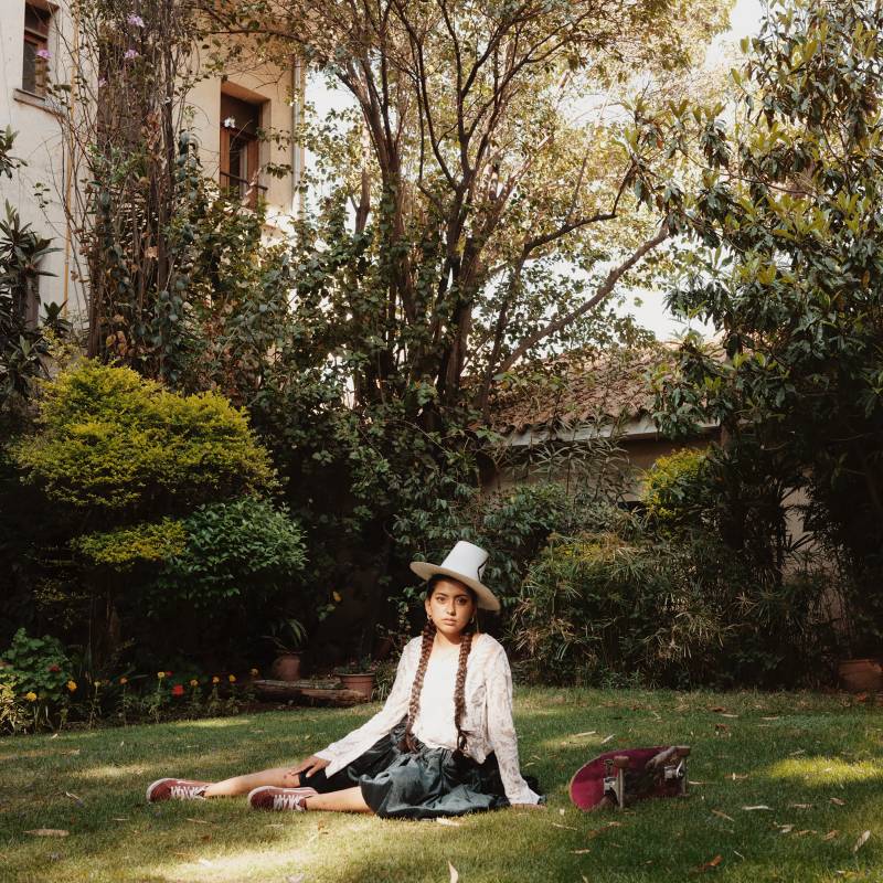 A young woman in traditional Bolivian dress and hat sits elegantly on a green lawn, surrounded by trees.
