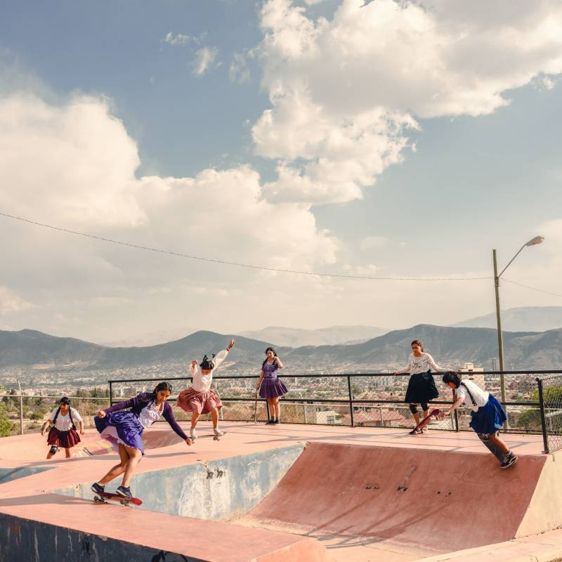 Six girls practice skateboarding on a ramp. Mountains and the city serve as a dramatic backdrop.