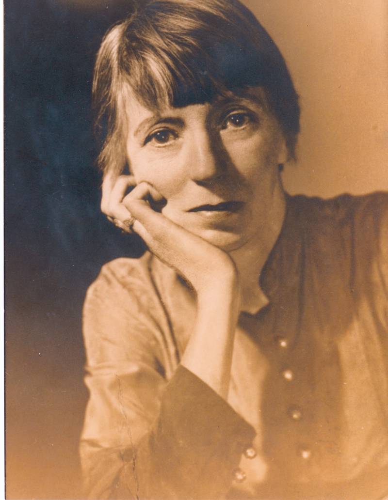 A sepia-toned photograph of a serious looking woman with sharp features, resting her chin in her hand.