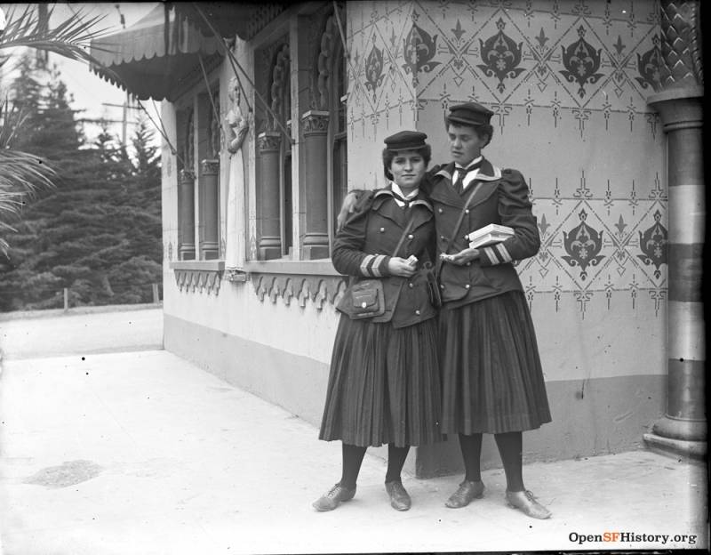 Two young women stand close together, the taller with her arm around the shorter. They wear matching uniforms of calve-length skirts, formal jackets, white shirts and ties, and petite caps.