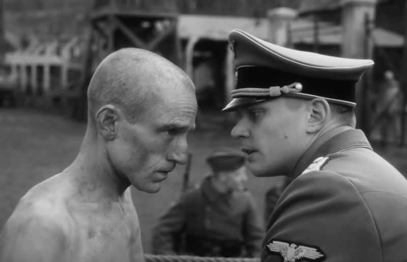 An emaciated shirtless man with a shaved head stands with a look of steely determination. Opposite a nazi SS officer gives him instructions. Behind them, fences and other soldiers are visible.