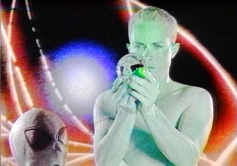 A green person in front of a psychedelic background holds a mysterious object.