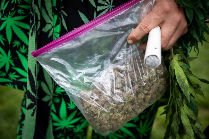 Close-up on a bag of marijuana, a large joint, held by a man in a marijuana-themed outfit 