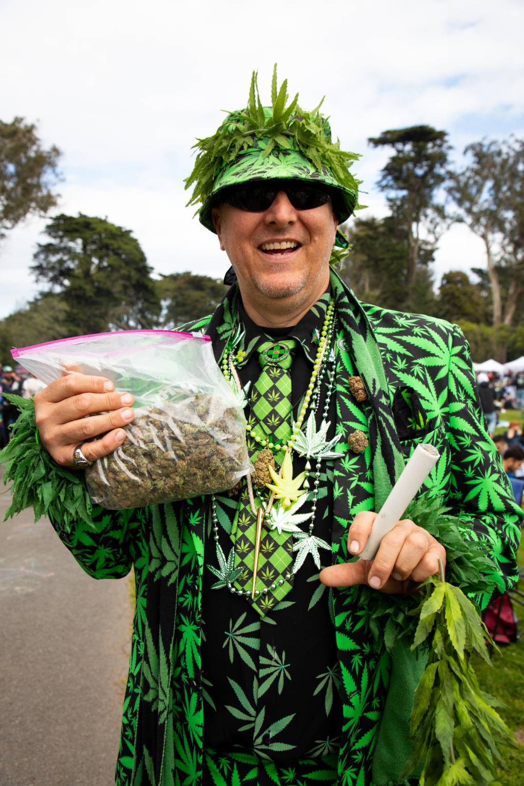 A middle-aged white man holds a bag of marijuana and a large joint, dressed in a marijuana-themed suit and bucket hat covered in leaves.