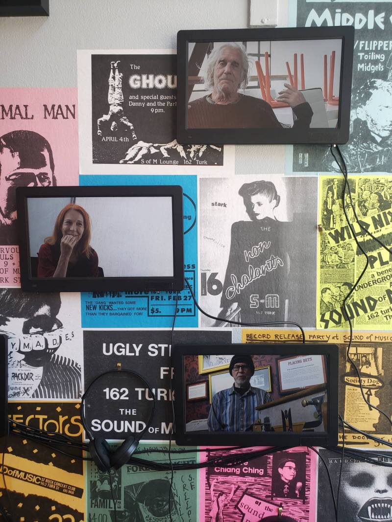 Three small flat screens mounted on a wall covered haphazardly with old punk flyers. Wires dangle from the screens.