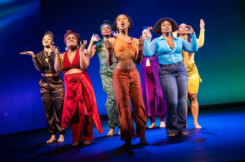 Seven women of color stands together in two rows, gesturing while singing. All are wearing brightly colored clothes.