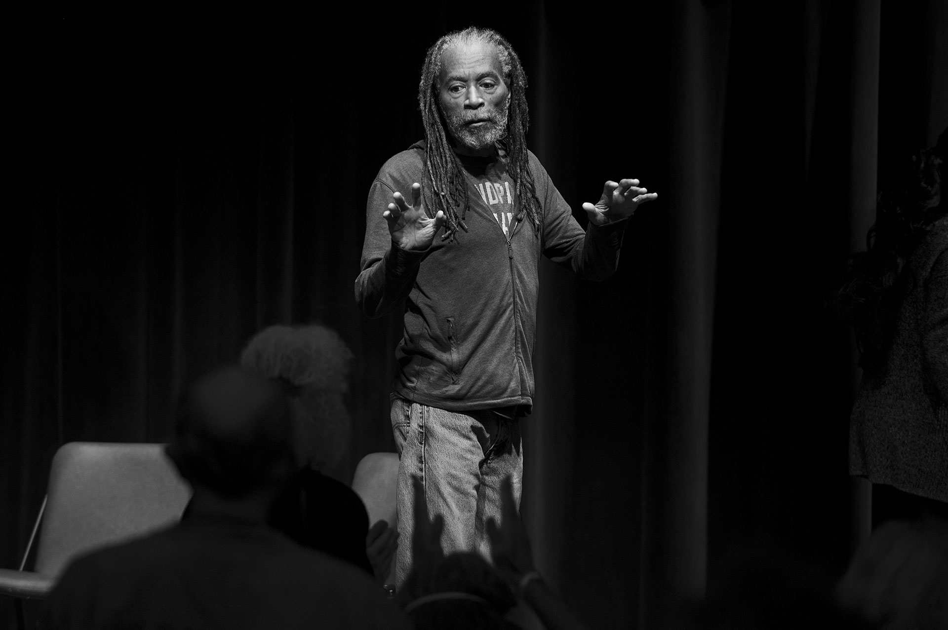An African American man in a sweatshirt, jeans, and dreadlocks on stage. 