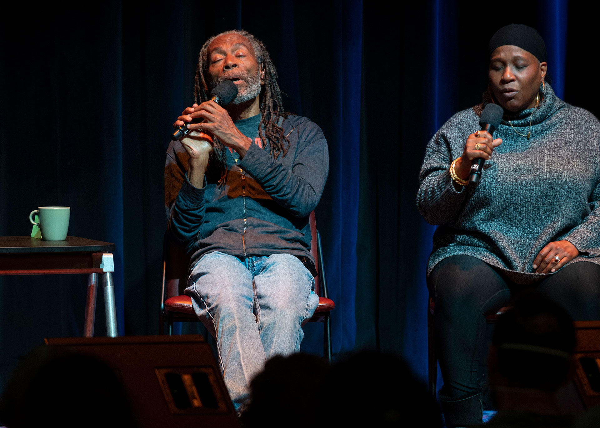 An African American man and woman sit on stage, singing into microphones