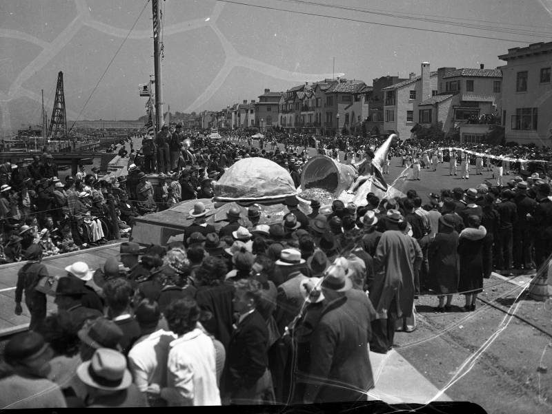 Crowds gather along a street of houses to watch a marching band, and a woman on a strange float driving past.