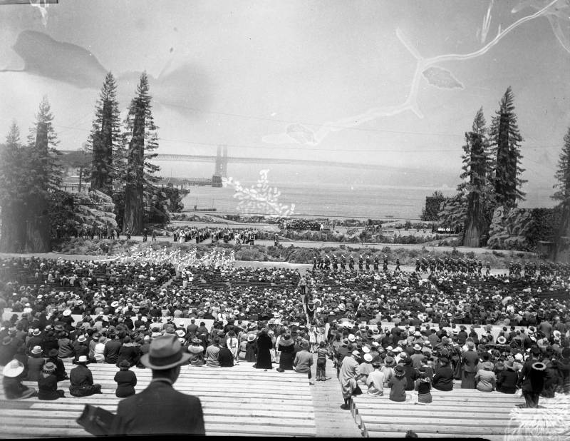 Row upon row of people gather on bleacher seats to watch marching bands. Further in the distance is the Golden Gate Bridge, most of which has been rendered invisible because of weather conditions.