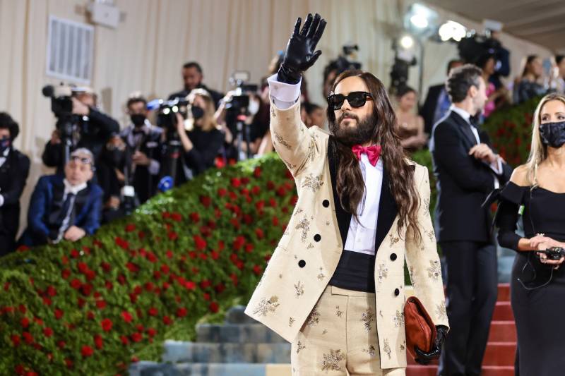 Jared Leto in a cream double breasted suit with black collar and button and red bow tie. He also wears dark sunglasses and black leather gloves.