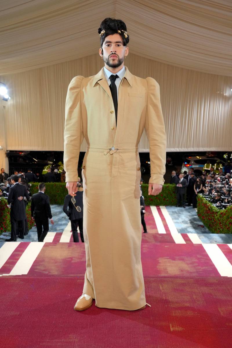 Bad Bunny in a slender, full-length beige coat with puffy sleeves and a white shirt and black tie underneath.