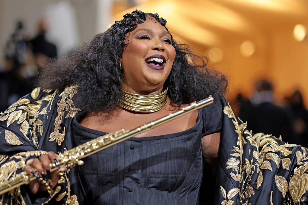 Lizzo smiles broadly, while wearing gold hoop necklaces, a black bodice and ornate black and gold coat. He is clutching a gold flute.