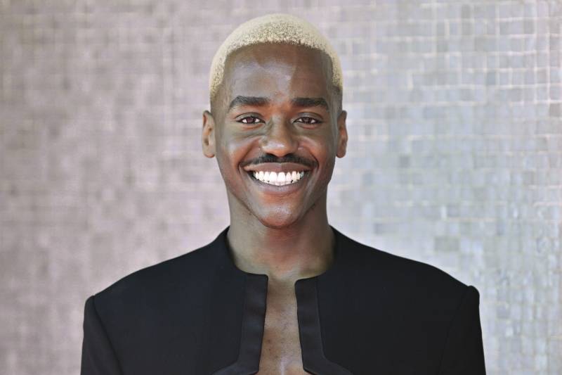A young Black man with tightly cropped bleached blond hair smiles broadly. He's wearing a black jacket with a few inches of his chest exposed.