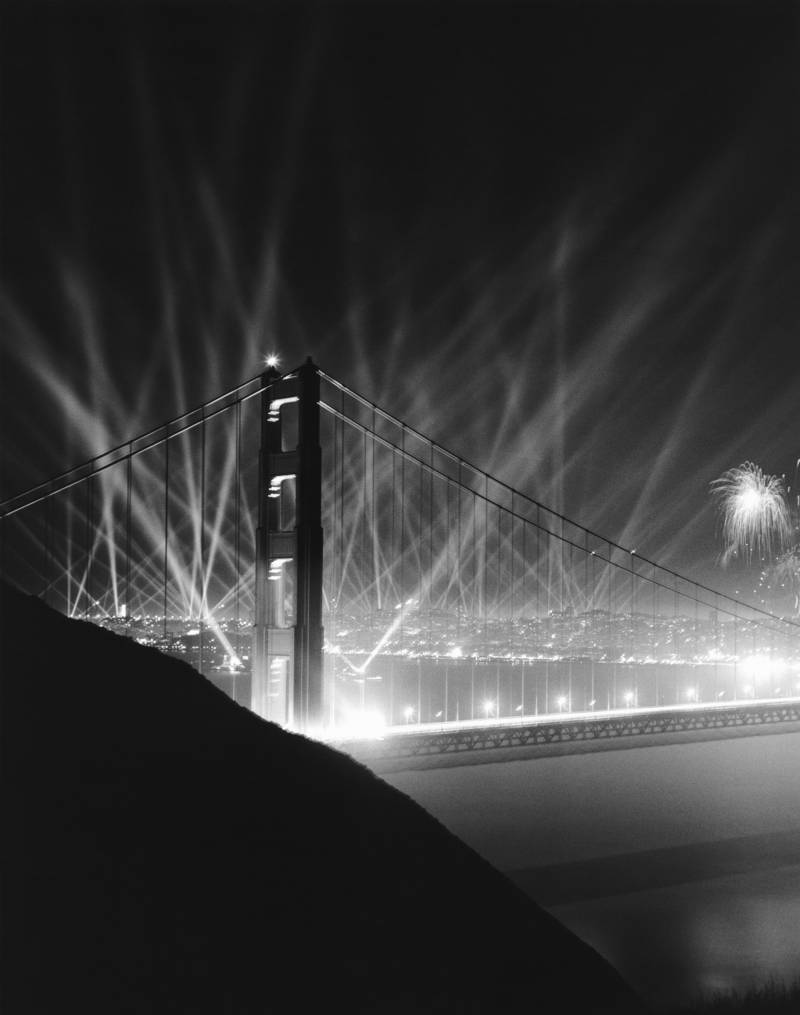 The Golden Gate Bridge, photographed in black and white, lit up by spotlights and fireworks.