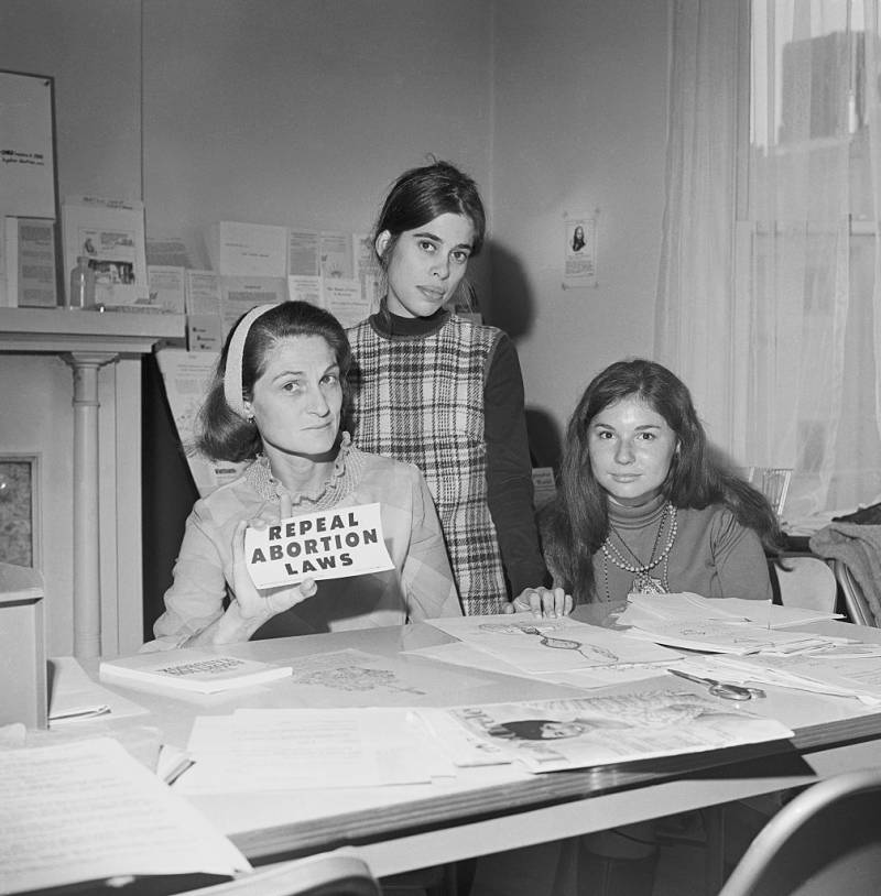 Three women wearing 1960s attire sit at a table strewn with papers. One of them holds up a sign that says 'REPEAL ABORTION LAWS'