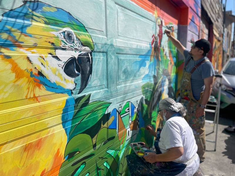 The image of a multicolored tropical bird, one of Josué Rojas' staple images, looks on as Esther and Josué finish a mural in Balmy Alley. 