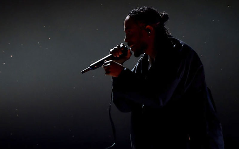 Kendrick Lamar, a young Black man, raps into a microphone in silhouette in the dark.