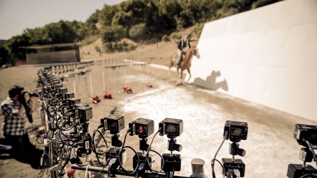 Rider and horse gallop in front of white wall faced by dozens of cameras