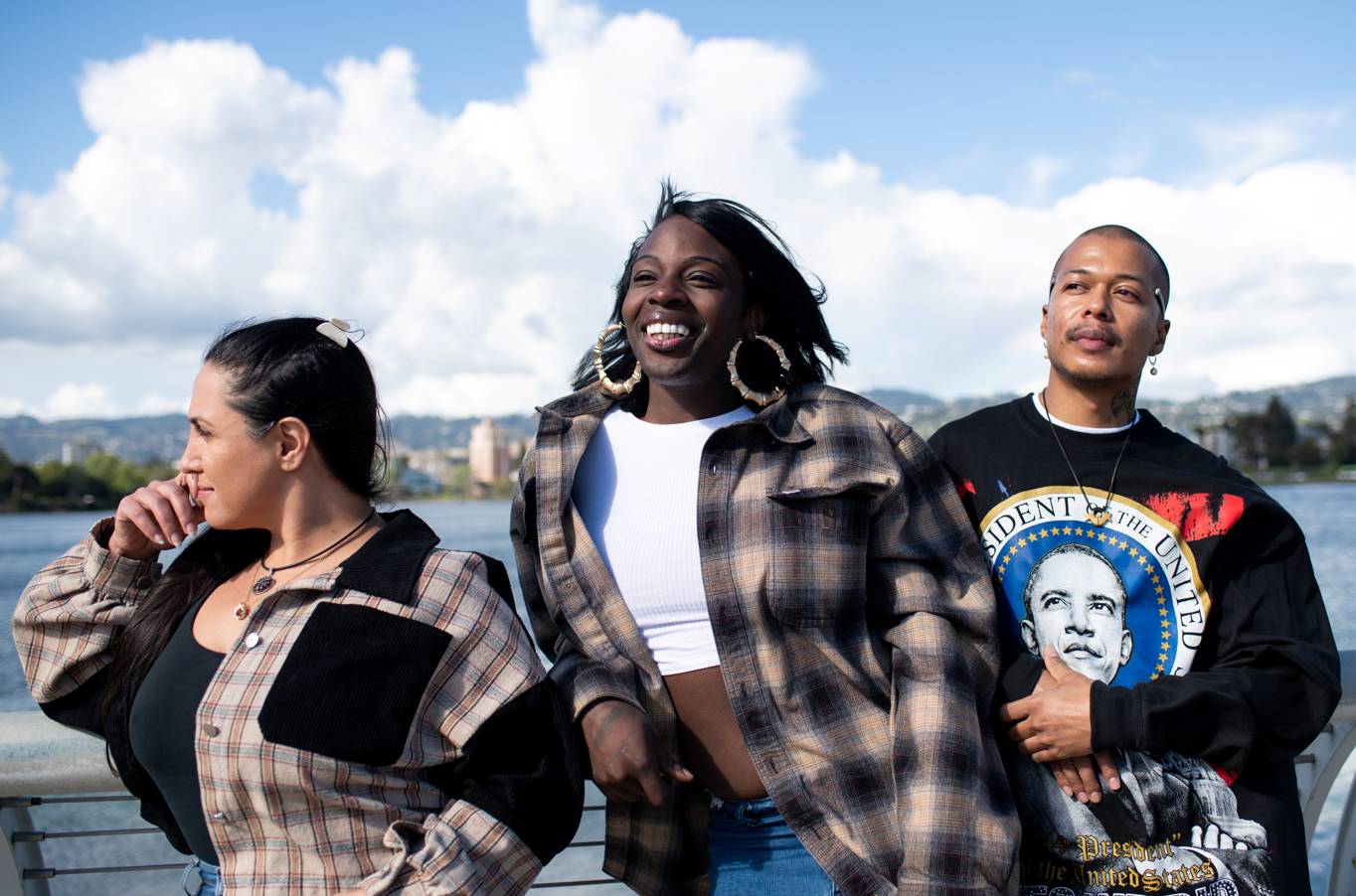Three people stand in front of Lake Merritt in Oakland and pose while looking away from the camera, to their right.