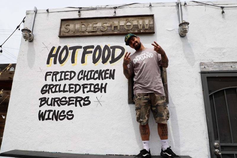 Man holds up two peace signs while standing in front of his restaurant named Sideshow, with hand painted sign behind him that says hot food