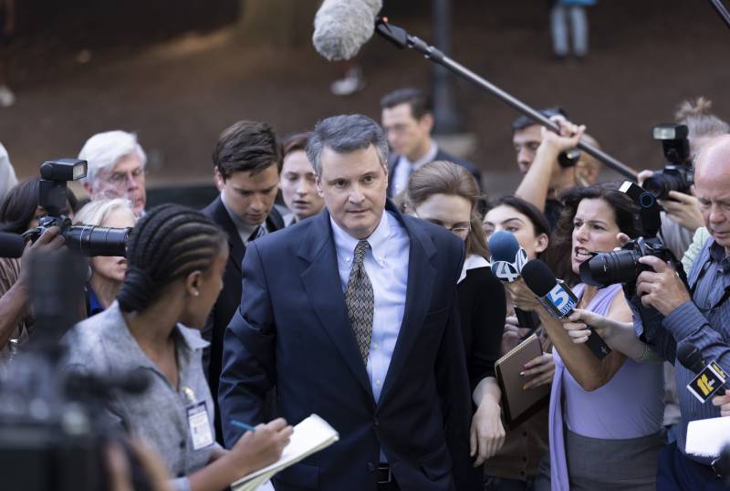 A white man wearing a suit walks through a tight crowd of reporters and media crews.