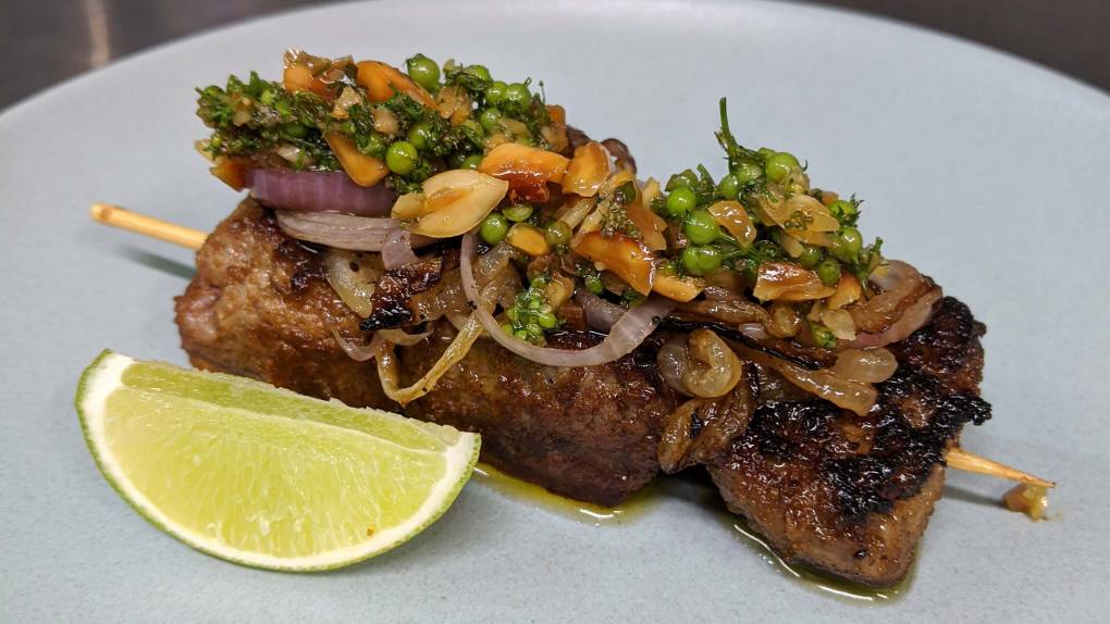 A grilled beef skewer topped with various herbs and spices, with a wedge of lime on the side.