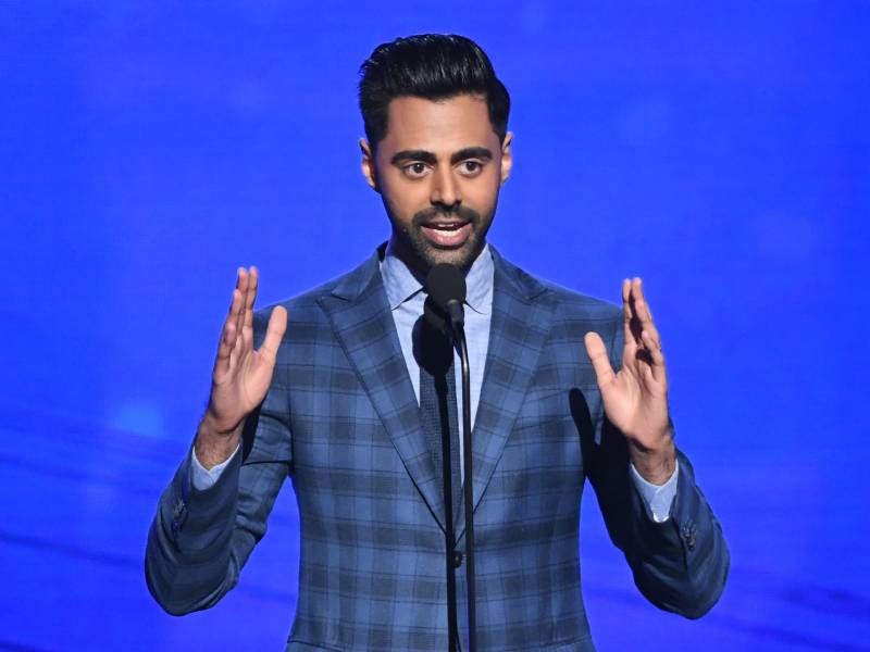 A man of Indian descent talks animatedly on stage before a blue backdrop. He wears a smart blue plaid suit and pale blue shirt.