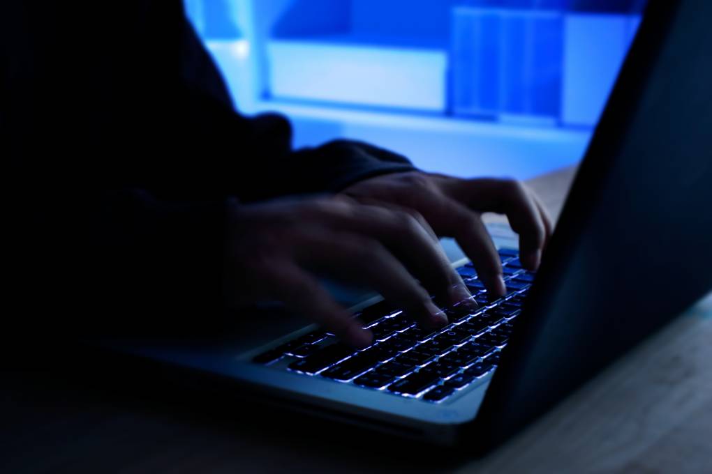Hands type on a laptop keyboard in a dimly lit room. A computer screen is visible behind the person typing.