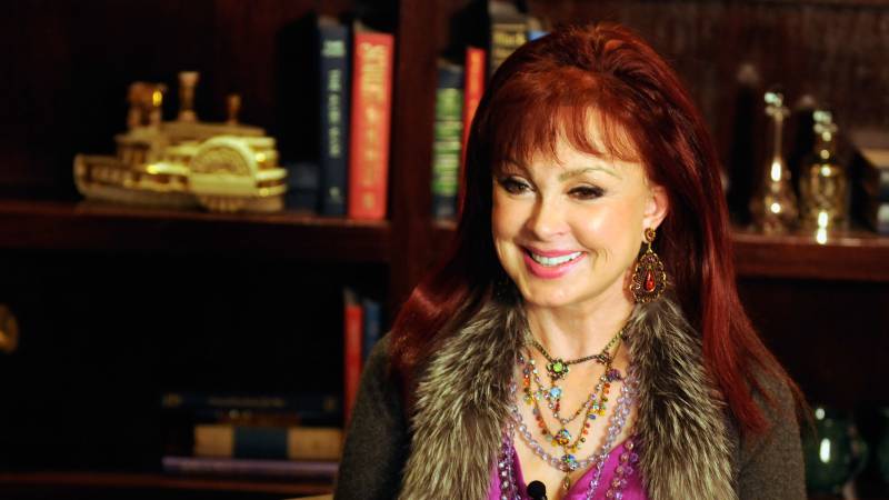 A vibrant-looking white woman with red hair smiles broadly , seated before a bookcase.