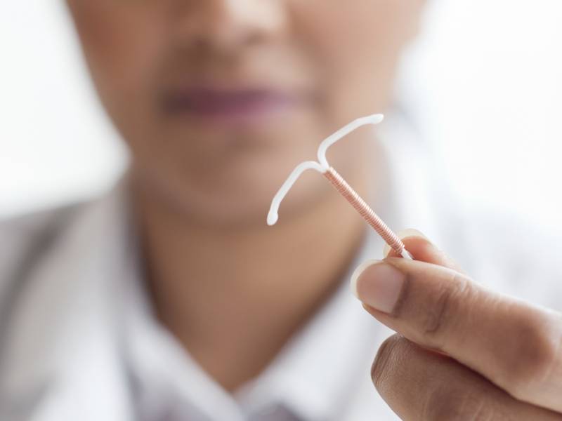 Close up of an IUD device, held between the fingers of a woman wearing a white shirt and lab coat.