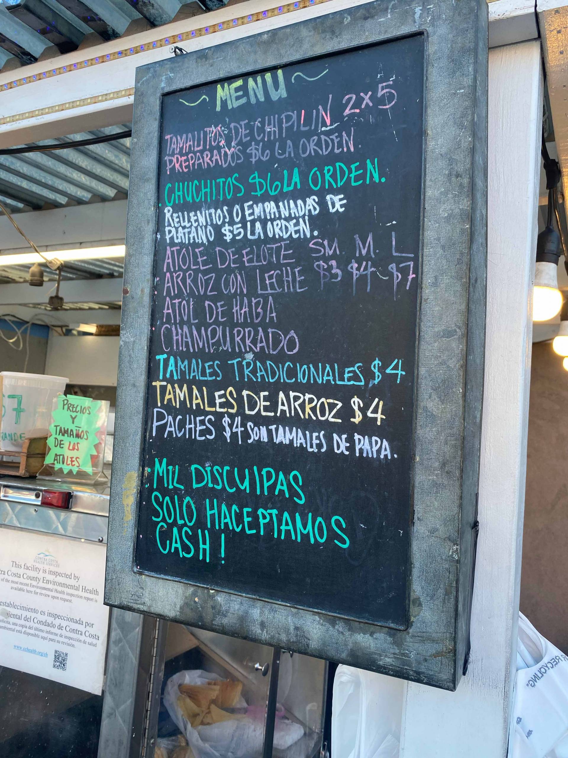 The menu signboard in front a food cart: Items include chuchitos, atol de elote, tamales tradicionales and tamales de arroz.