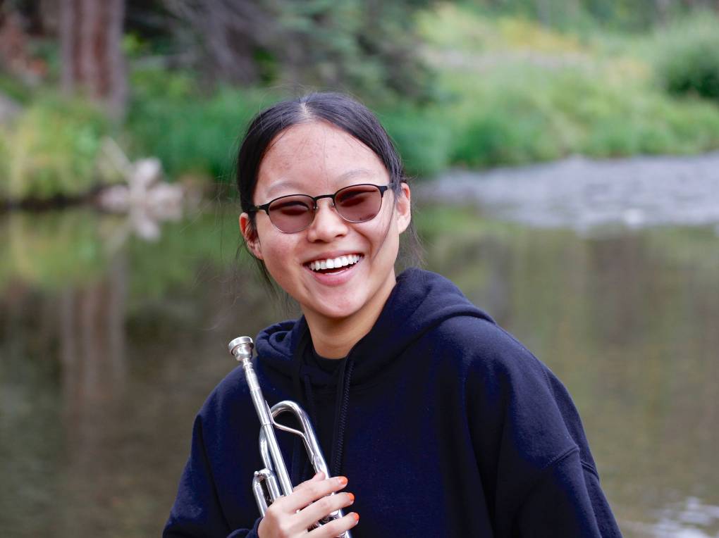 A young Asian woman wearing dark glasses and a black hoodie, stands smiling and clutching a trumpet close to her chest, in front of a lake.