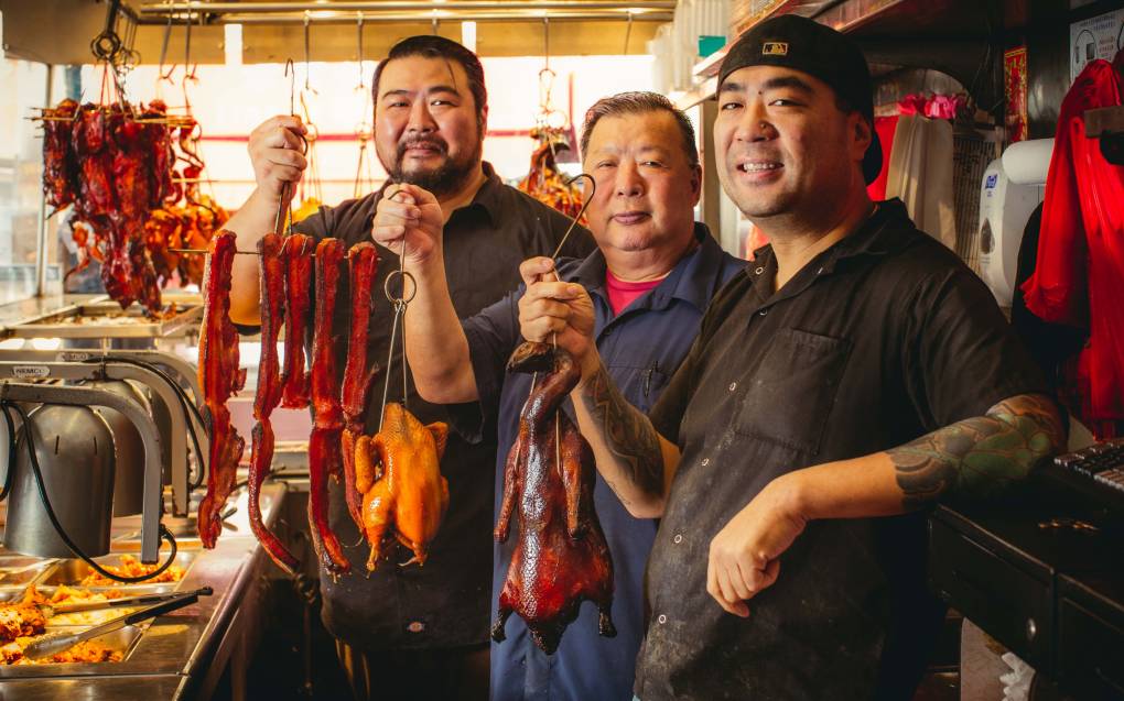 A father and two brothers—all Chinese American—hold up a selection of barbecued meats inside their restaurant.