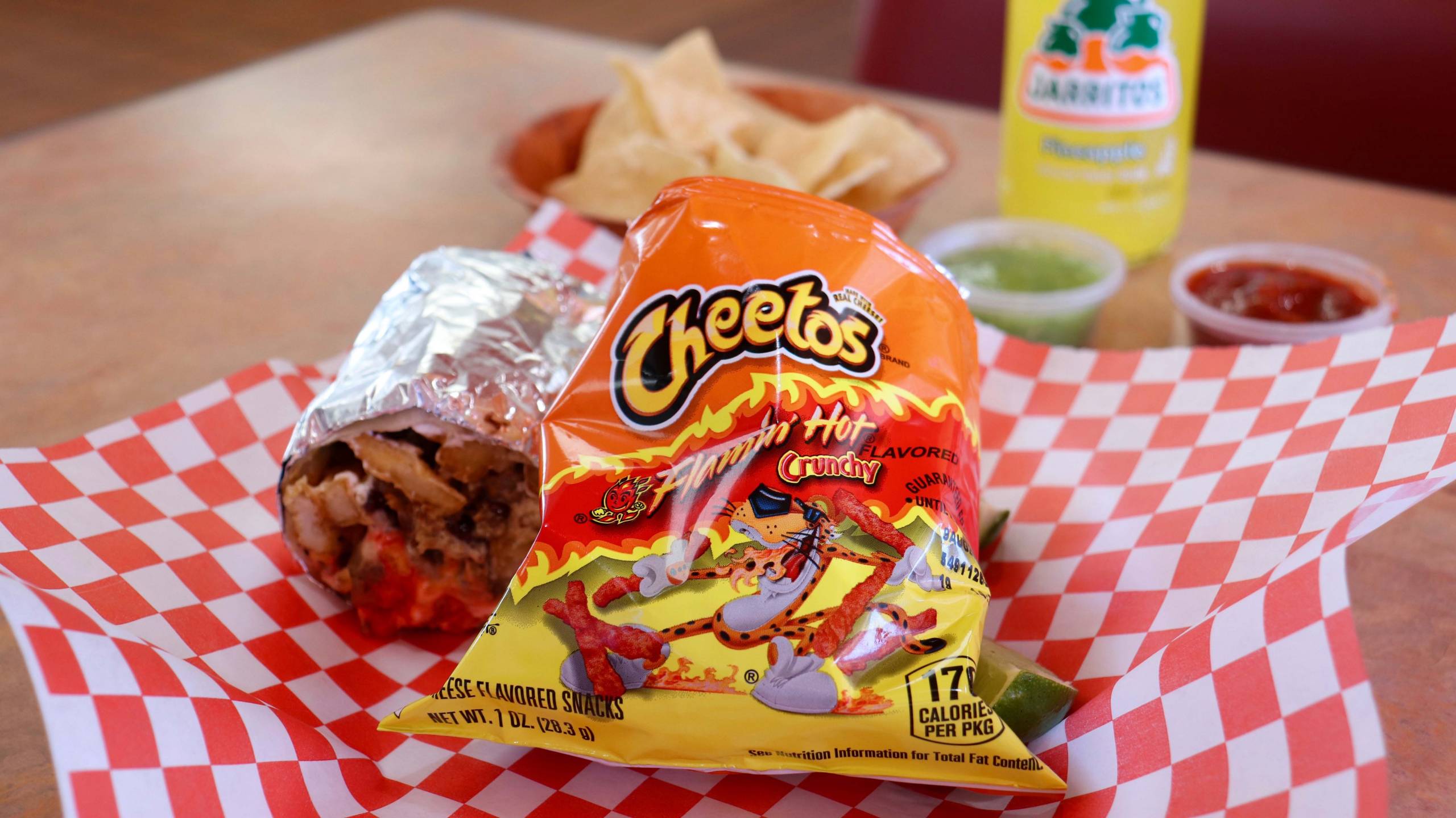 A foil-wrapped burrito and a bag of Flamin' Hot Cheetos on red and white checkered butcher paper.