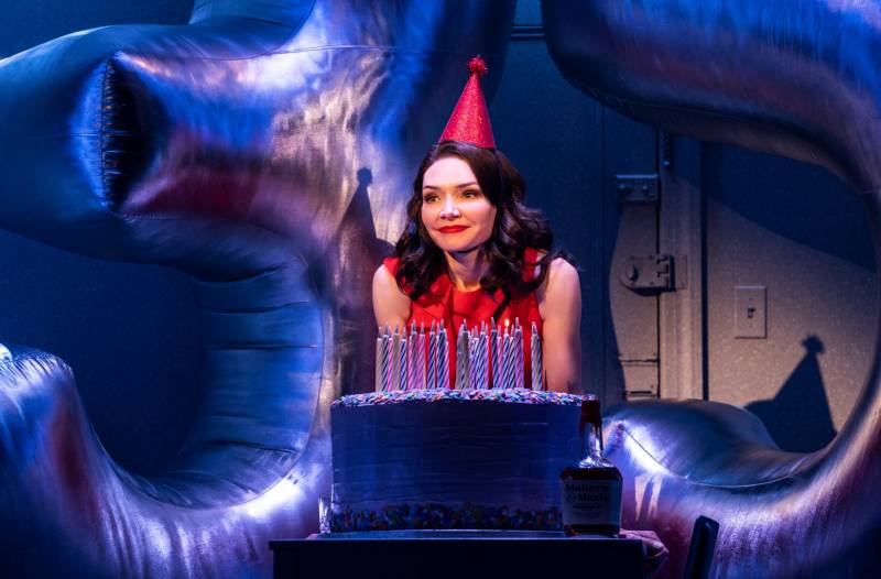 A young woman wearing a red dress and red party hat sits behind a large birthday cake, looking quietly happy.
