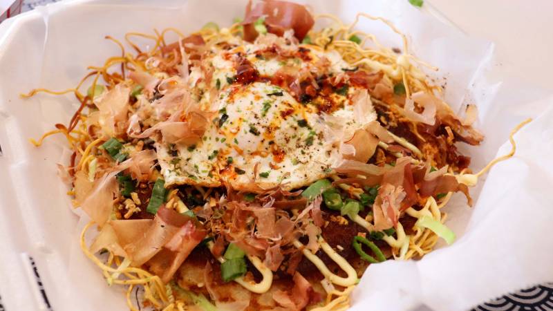 A plate of okonomiyaki, which is a Japanese style pancake. The ingredients include a fried egg, dried fish, mayonnaise, and scallions, all drizzled on top.