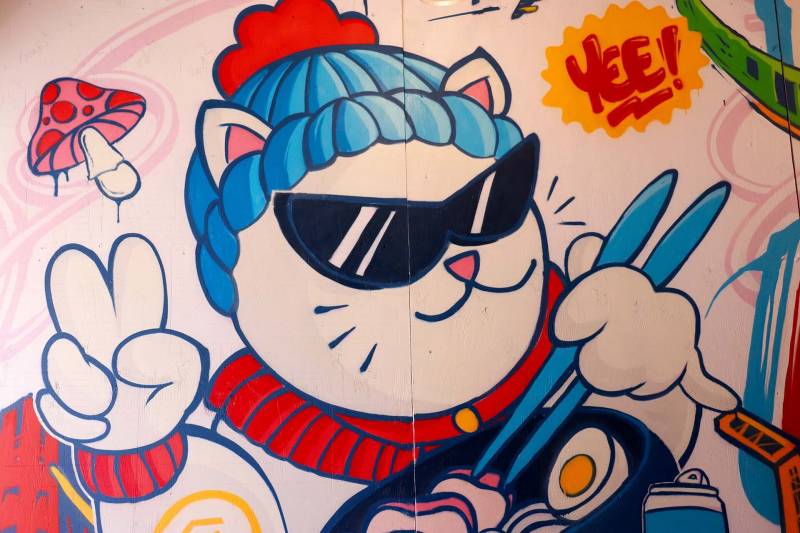 A cartoon mural of an Asian cat wearing a beanie, sunglasses, and holding chopsticks. The mural is located inside Baba's House Kitchen restuarant.