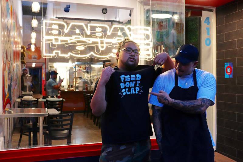 Two young men, Paris and Joog, stand in front of Baba's House Kitchen. Joog is wearing a shirt that says "food don't slap" while P is beside him, pointing at the shirt.