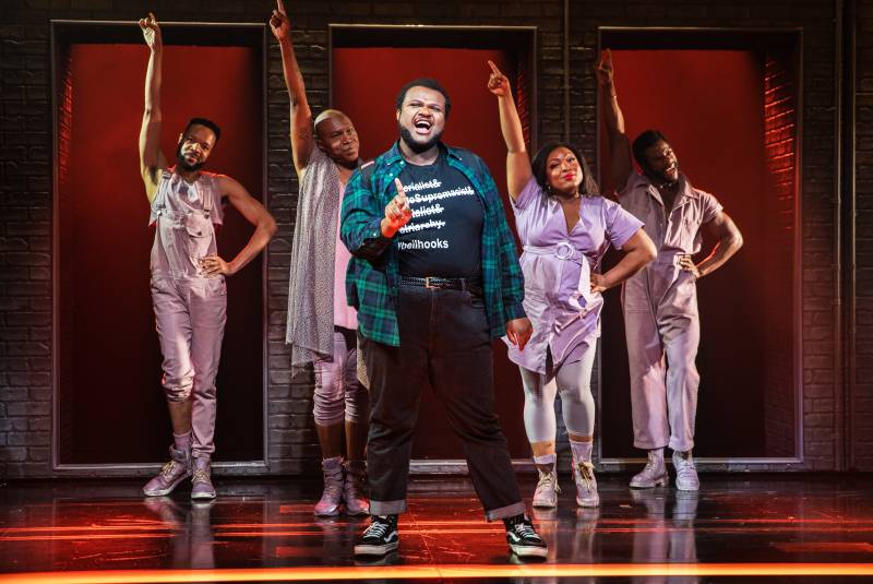 A black man wearing jeans, T-shirt and casual flannel stands singing, finger pointed up to the ceiling. Behind him are four dancers wearing lilac and striking a theatrical pose.