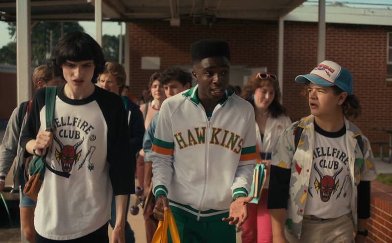 A Black teenage boy walks, chatting animatedly between two white friends. He is wearing an athletic jacket that says 'HAWKINS' on the front. They are wearing T-shirts with devil emblems on them that read "Hellfire Club."