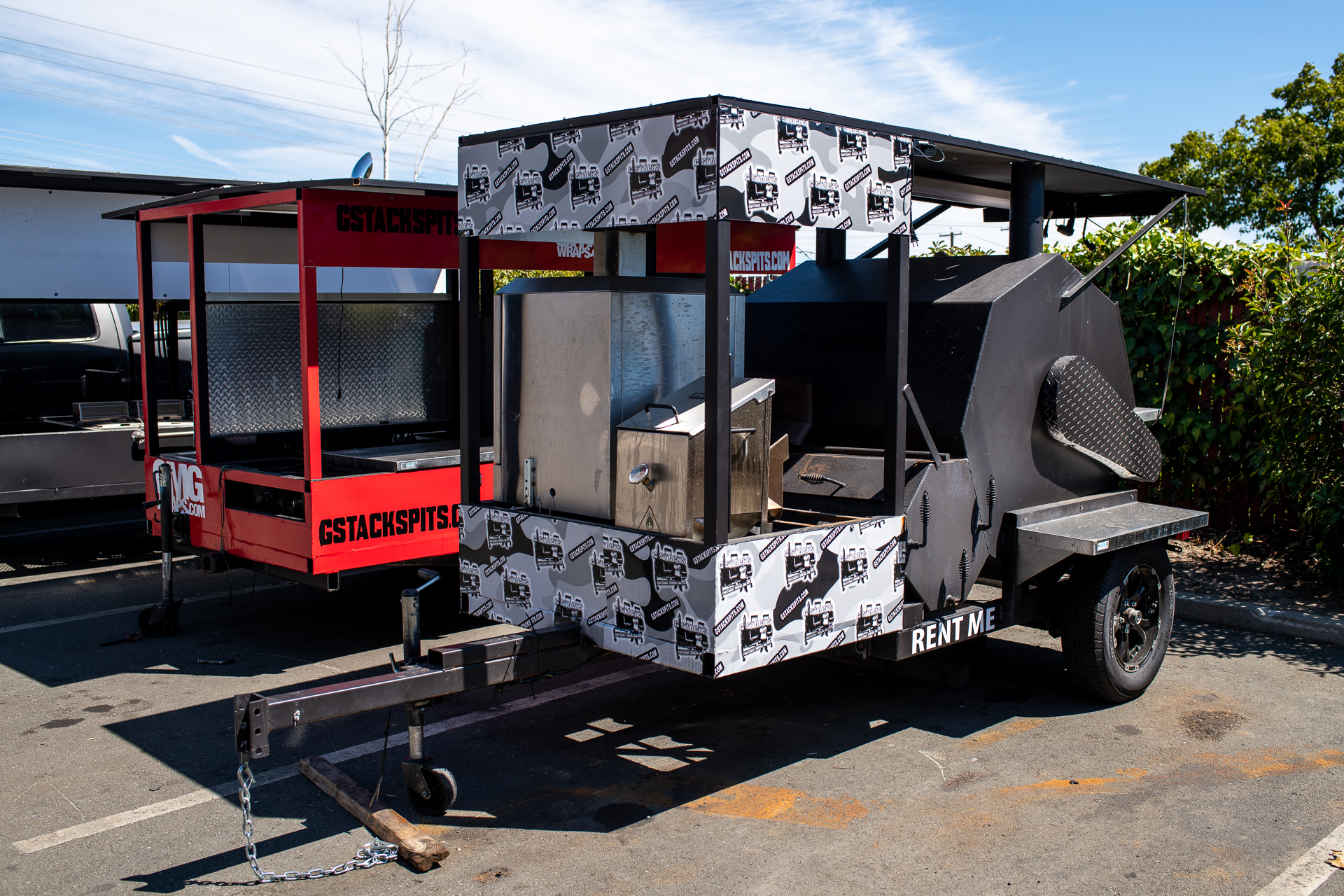 A black and white barbecue smoker on wheels in an empty lot; a red smoker is behind it.